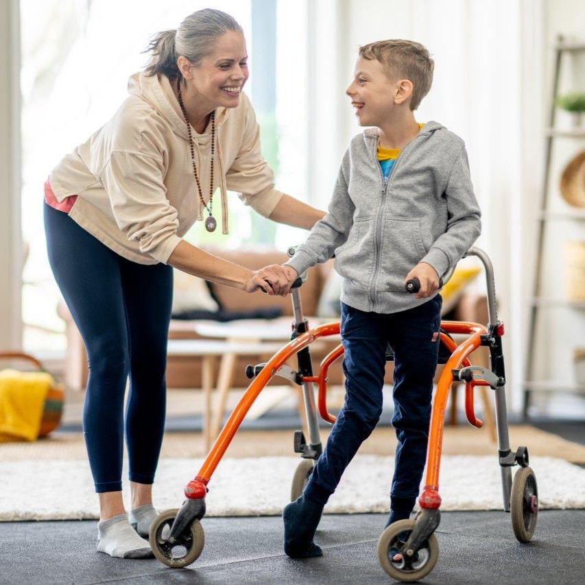 A woman supports a young boy who is moving forward with the help of a rollator.