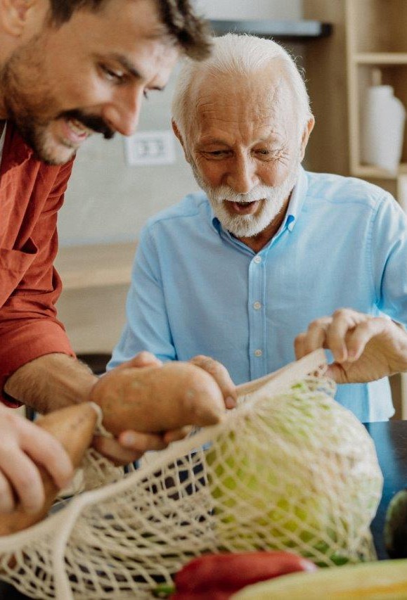 A younger man unpacks vegetables from a shopping bag. Next to him sits an older man who is smiling as he looks at the groceries.